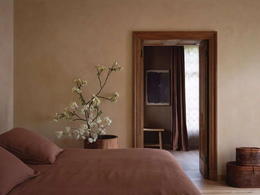 Bedroom interior featuring a bed with brown linens, a flowering branch in a pot, and a doorway leading to another room.
