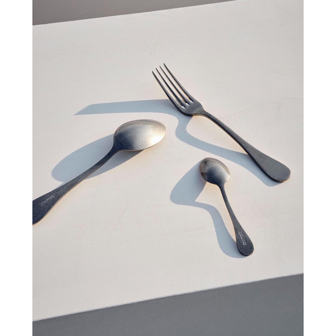 Silverware set including a fork, a large spoon, and a small spoon arranged on a white surface with natural lighting creating shadows.