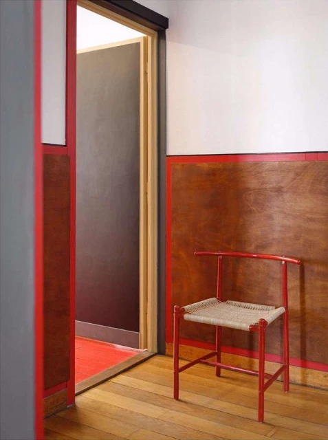 Interior view of a red painted chair with a woven seat on a wood floor with a doorway leading to another room with a red floor.