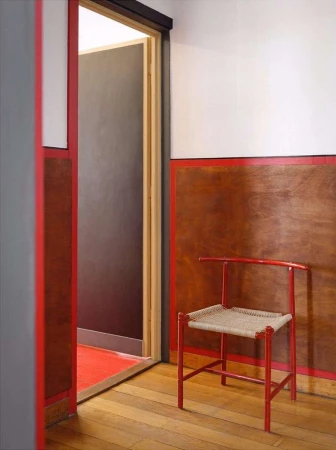 Interior view of a red painted chair with a woven seat on a wood floor with a doorway leading to another room with a red floor.