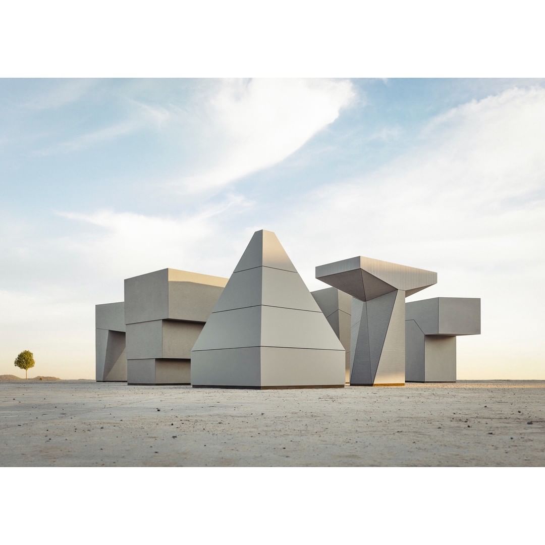 Geometric architectural forms stand in a barren landscape under a blue sky, with a single tree visible in the distance. 