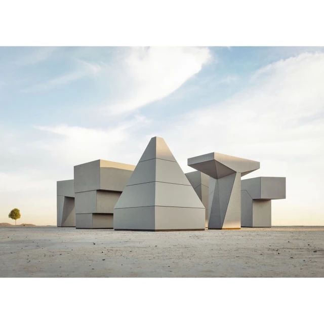 Geometric architectural forms stand in a barren landscape under a blue sky, with a single tree visible in the distance. 