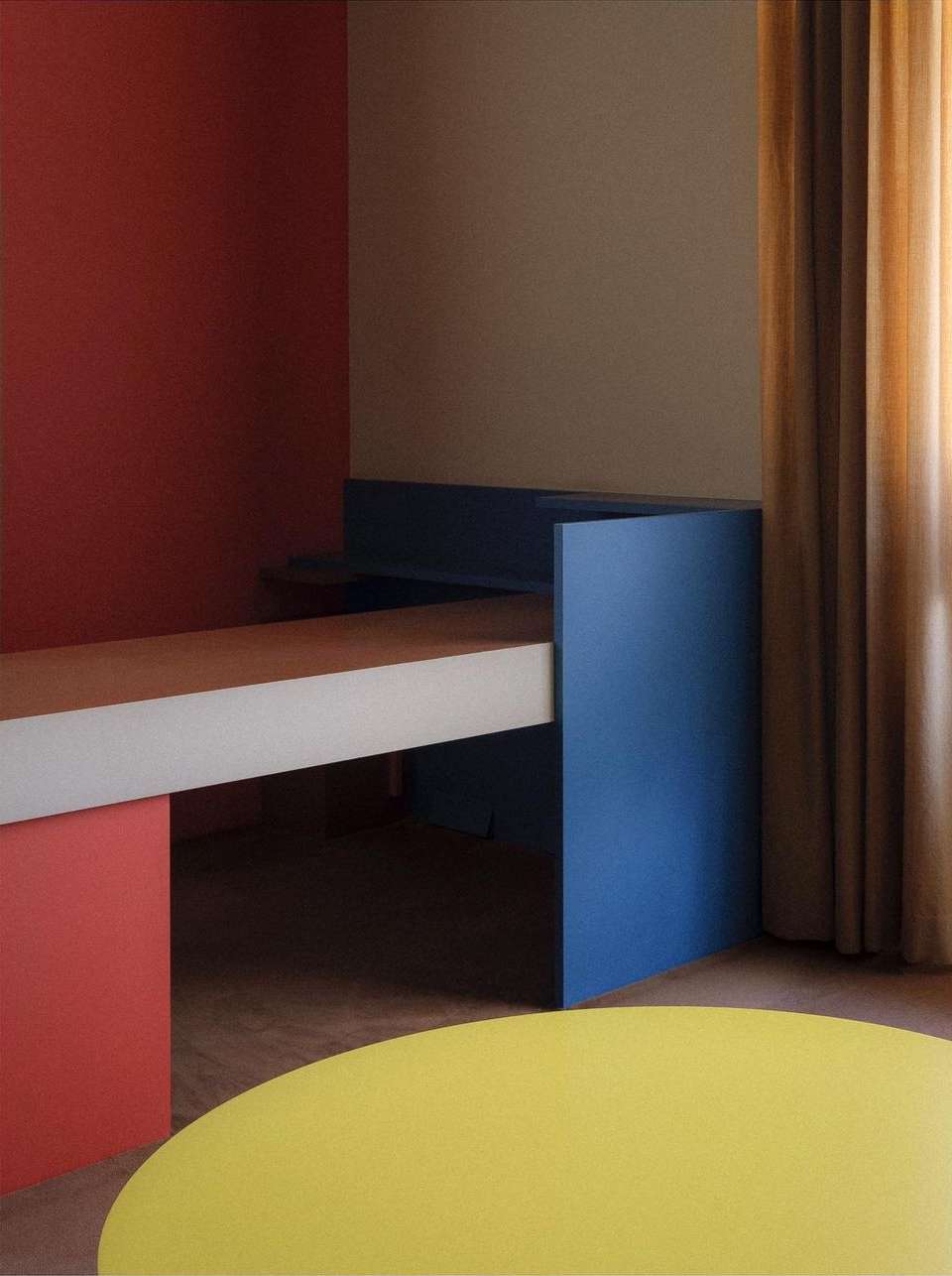 Geometric interior with blue desk, yellow rug, and red wall, creating a colorful, minimalist design. Features natural light and flat colors.