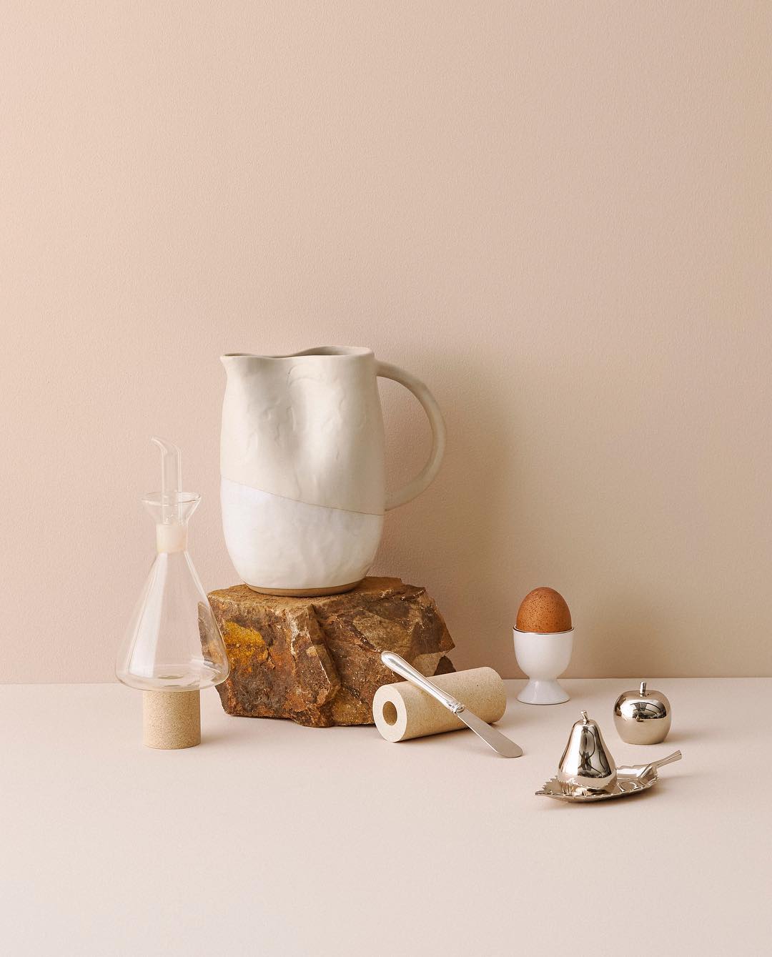 Still life featuring a ceramic pitcher on a stone, with a glass decanter, brown egg, and silver serving pieces on a pale surface.
