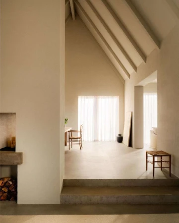 Minimalist interior with high vaulted ceiling, exposed wooden beams, pale neutral walls, and a recessed fireplace with stacked logs.