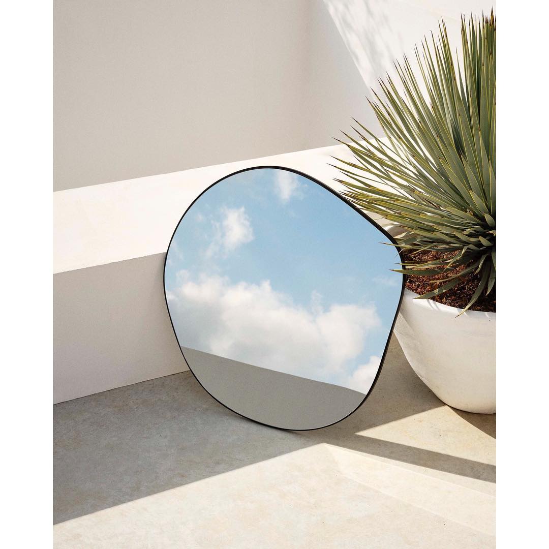 Irregular-shaped mirror reflects a sky with clouds, positioned near a potted plant against a white wall and concrete floor, creating a modern interior.