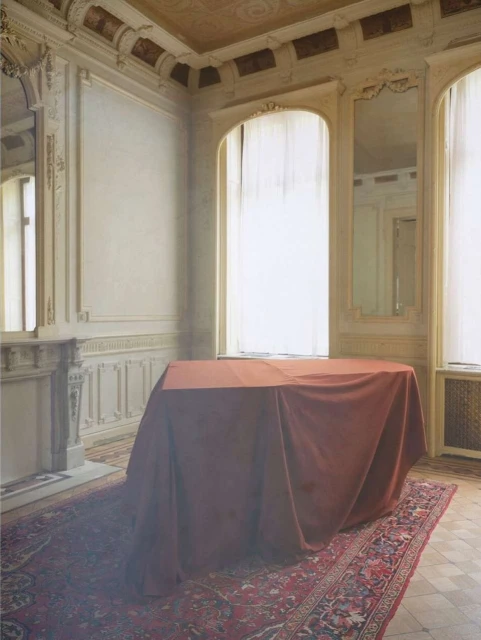 Ornate room with a large table draped in red fabric, illuminated by natural light streaming through a sheer-curtained arched window.