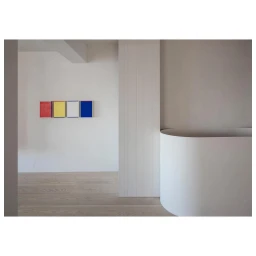 Minimalist interior with light wood flooring, white walls, and a row of four colorful framed artworks mounted on the wall.