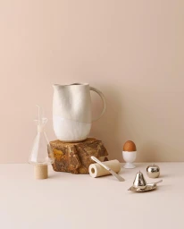 Ceramic pitcher on rock fragment with glass cruet, small egg in cup, and metallic fruit ornaments on pale backdrop