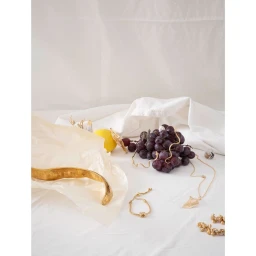 Still life photography featuring a bunch of dark grapes, a yellow lemon, and gold jewellery arranged on draped white fabric.