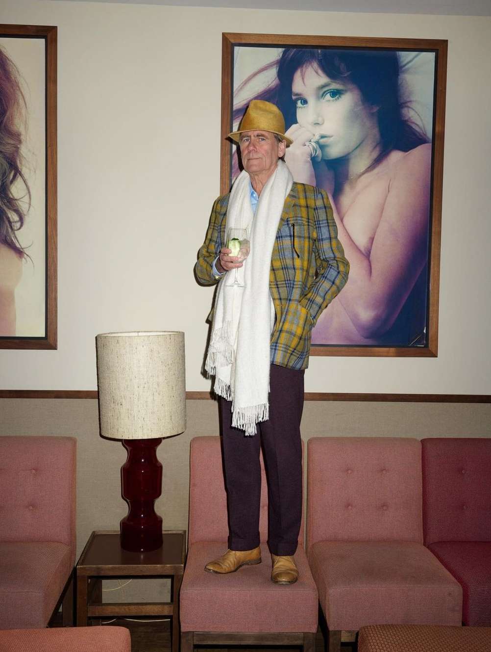 Elderly man in plaid jacket and hat stands on pink chairs indoors, holding drink. Large framed photograph hangs on the wall behind.