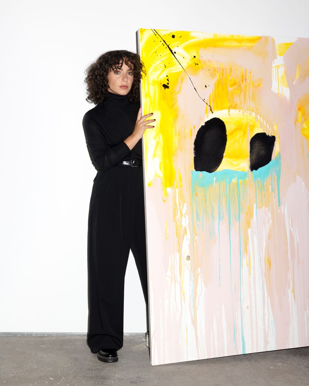 An adult woman in a black turtleneck and pants stands next to a large abstract painting with pastel colors and bold black shapes.