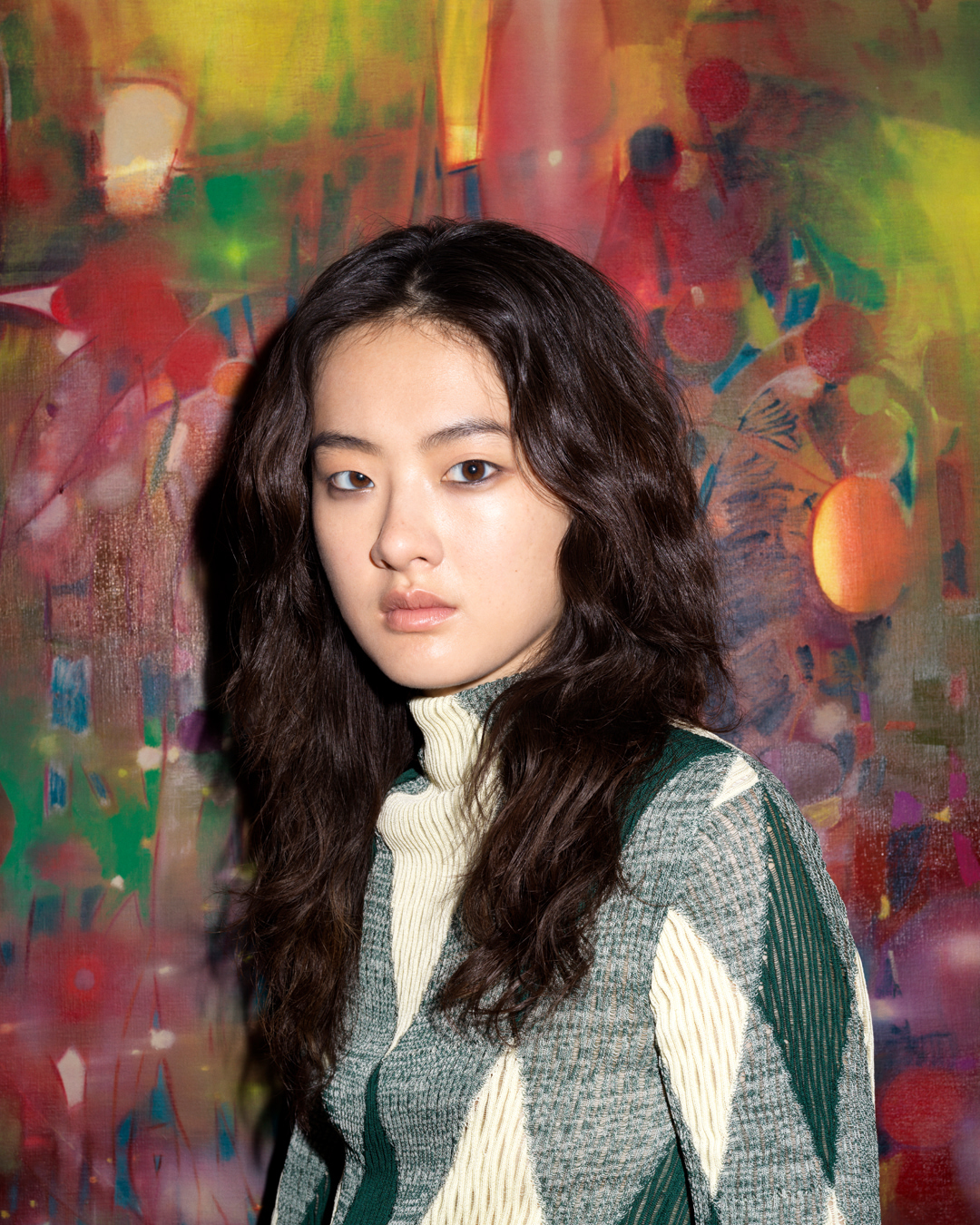 Portrait of a young woman with dark wavy hair wearing a patterned turtleneck sweater against a colorful painted background.