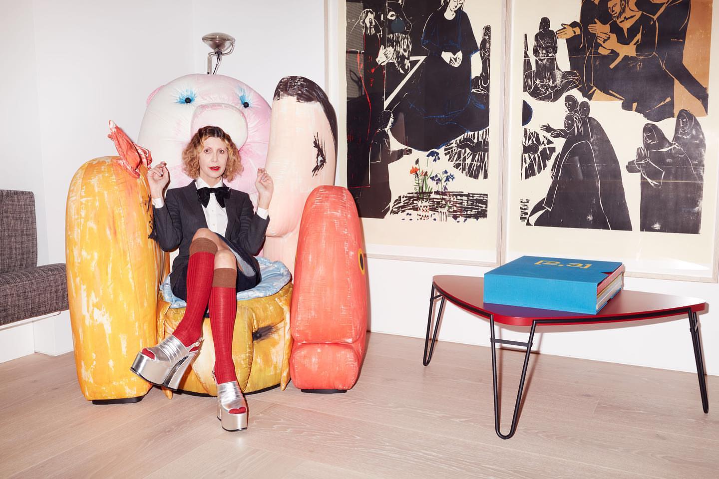 Elegant adult woman with curly hair, seated amidst colorful objects in a light-filled interior setting, wearing silver platform shoes.