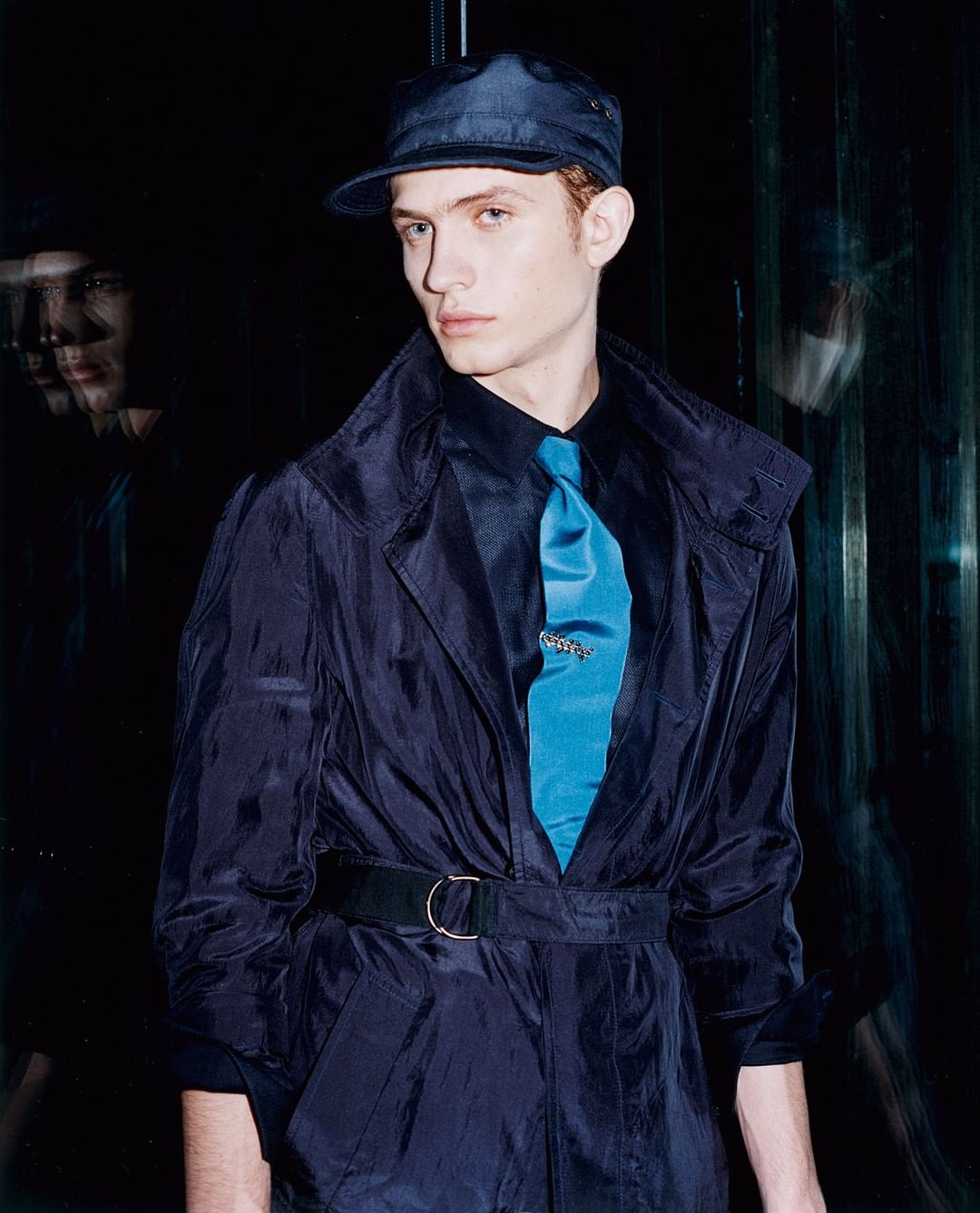 Stylish portrait of a young adult man in a navy coat, cap, and turquoise tie, standing against a dark background with blurred reflections.