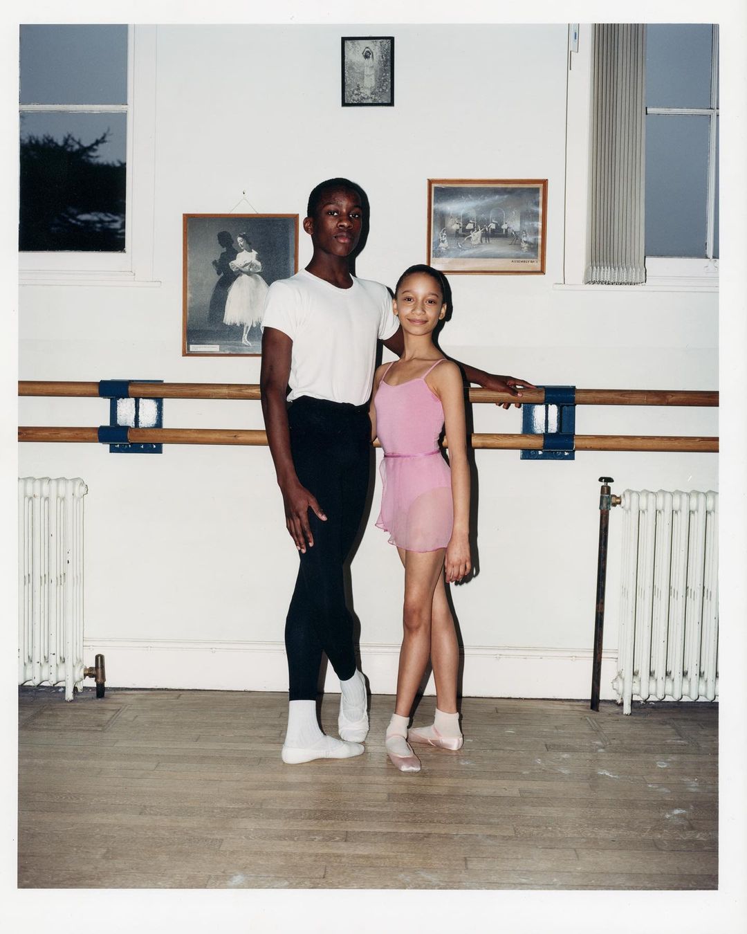 Two young ballet dancers, an adult male and a teenage female, pose in a dance studio with ballet barre, wooden floor, and natural light.