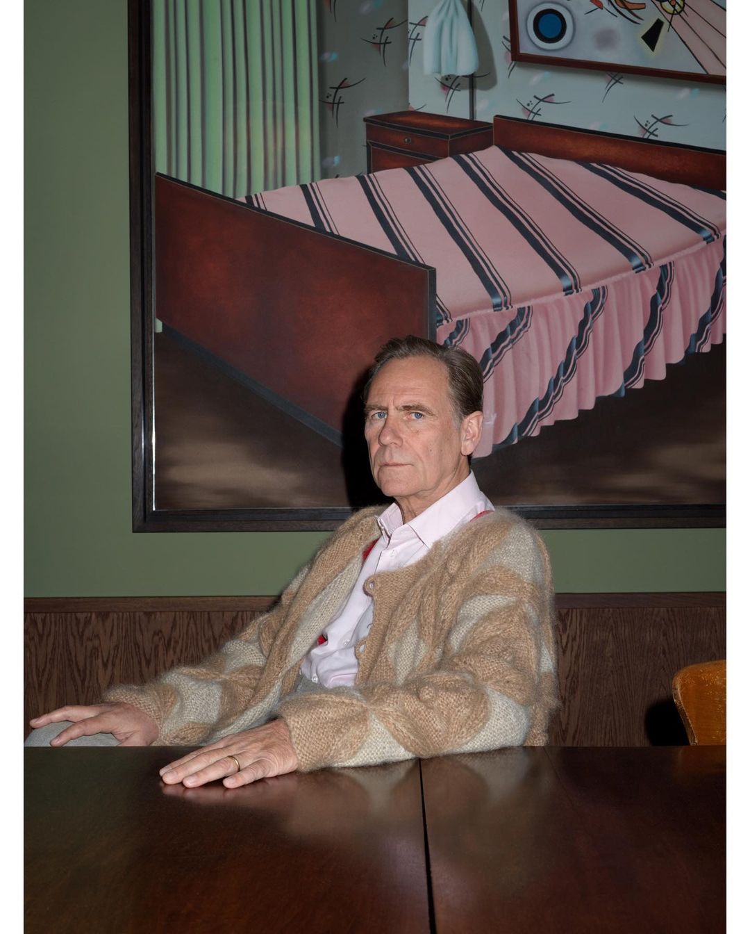 Elderly man with tan cardigan sits at a table in front of a framed painting of a bedroom. Indoor portrait with muted tones.
