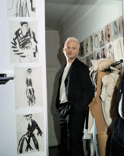 Indoor portrait of a fair-skinned adult man with short blonde hair wearing a black suit jacket, standing near fashion sketches and clothing racks.