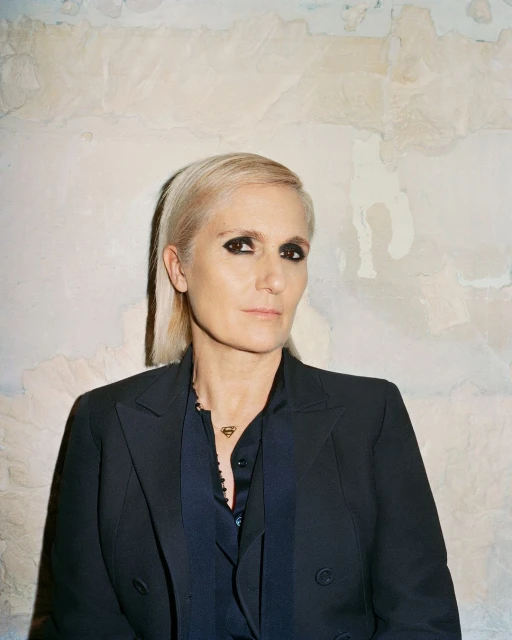 Portrait of fair-skinned, light-haired adult female in dark blazer and dark button-down shirt, wearing Superman logo necklace, against textured wall.