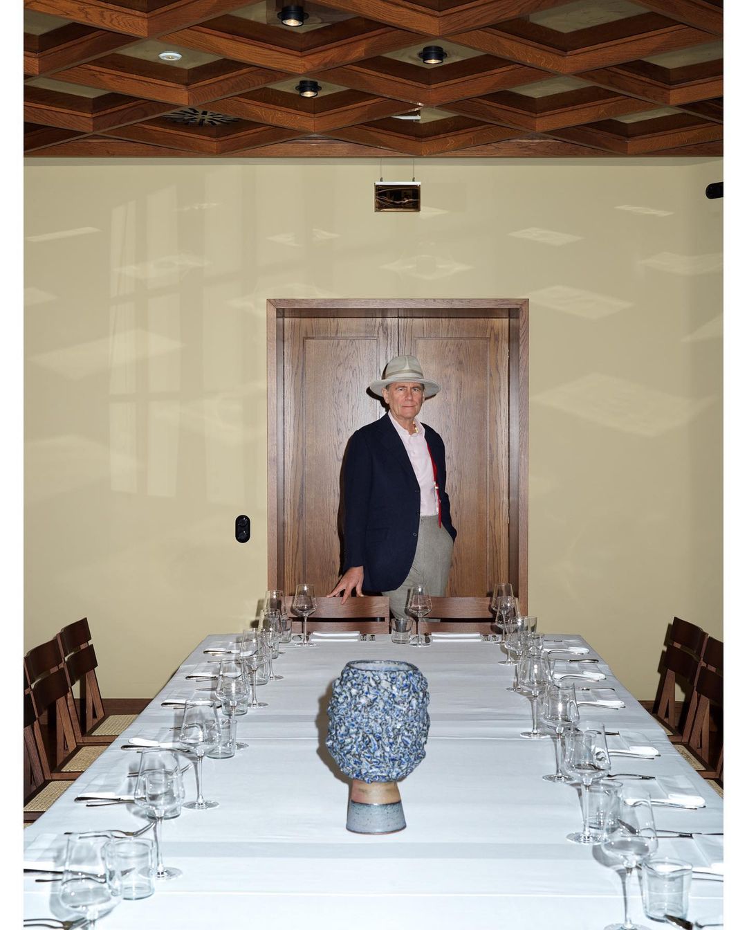 An adult man wearing a hat stands at the head of a long dining table set with glasses and flatware, in a wood-paneled room.