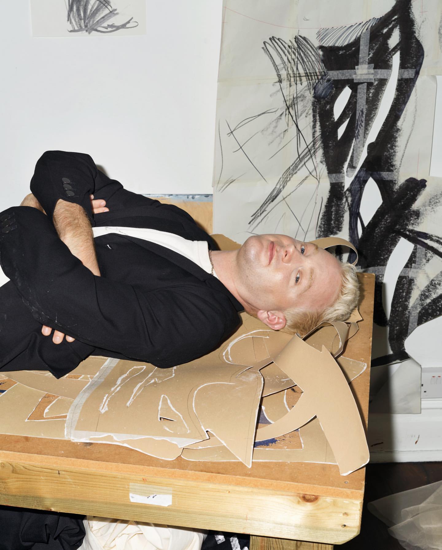 Adult male lying on a table covered with cardboard cutouts in an indoor environment surrounded by abstract drawings and art pieces.