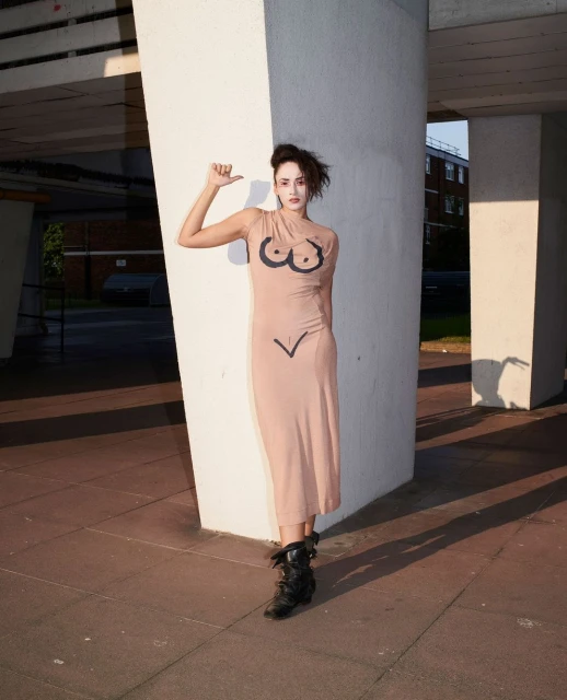 Adult woman poses outdoors in a beige dress with black body-like shapes, complemented by black boots, against a concrete building backdrop.