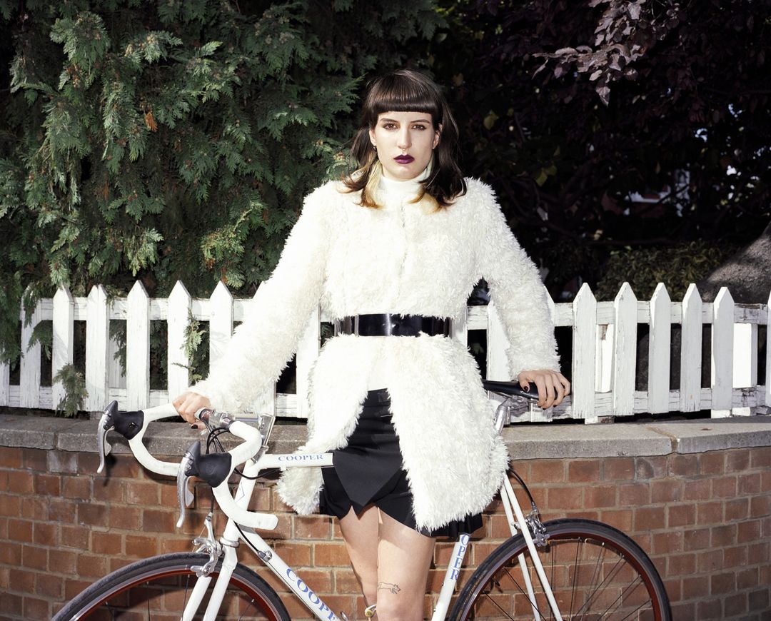 An adult female posing with a road bicycle in an urban environment with trees and a white picket fence, wearing a fuzzy white coat.