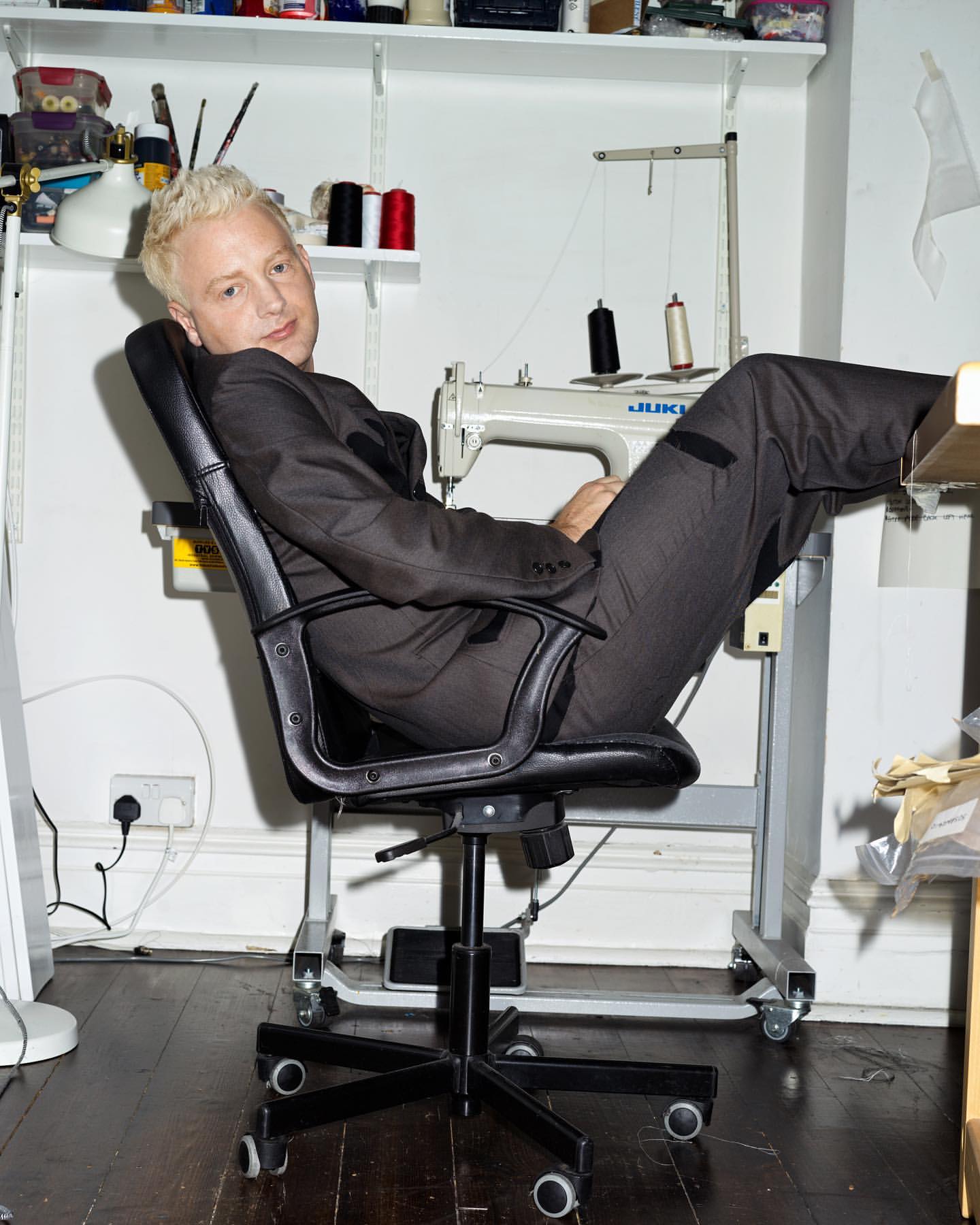 Blonde man in a grey suit sits in a black office chair in front of a sewing machine and assorted sewing supplies indoors.