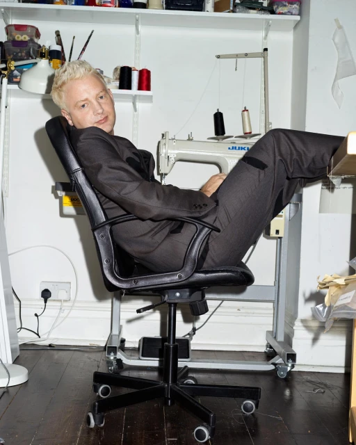 Blonde man in a grey suit sits in a black office chair in front of a sewing machine and assorted sewing supplies indoors.