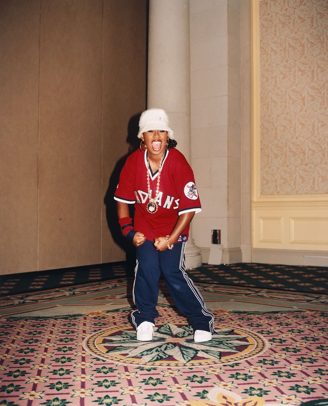An energetic adult female in a baseball jersey and bucket hat poses indoors on a patterned carpet, showcasing a dynamic and joyful presence.