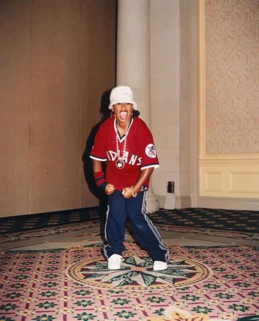 An energetic adult female in a baseball jersey and bucket hat poses indoors on a patterned carpet, showcasing a dynamic and joyful presence.