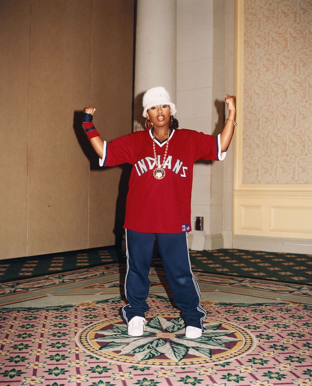 Adult woman in a red Indians baseball jersey, white bucket hat, and blue pants poses indoors with arms raised in a confident gesture.