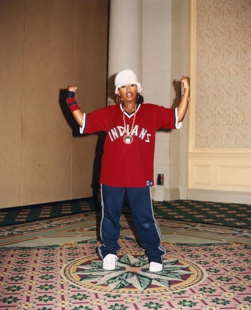 Adult woman in a red Indians baseball jersey, white bucket hat, and blue pants poses indoors with arms raised in a confident gesture.