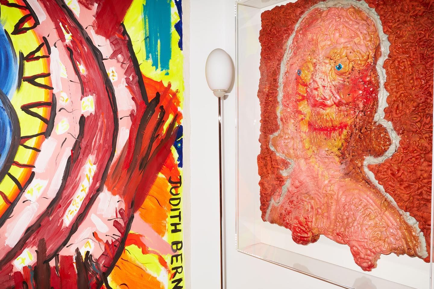 Two paintings on a white wall, one colorful and abstract, the other a red textured portrait under a floor lamp.