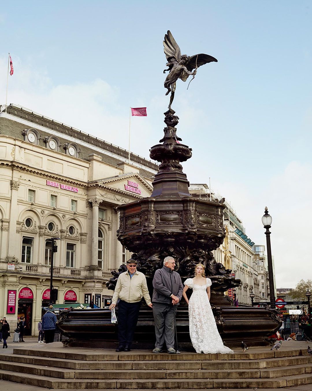 Two older men and a young woman in a white dress stand together on the steps of the Eros statue at Piccadilly Circus in London on a bright day.