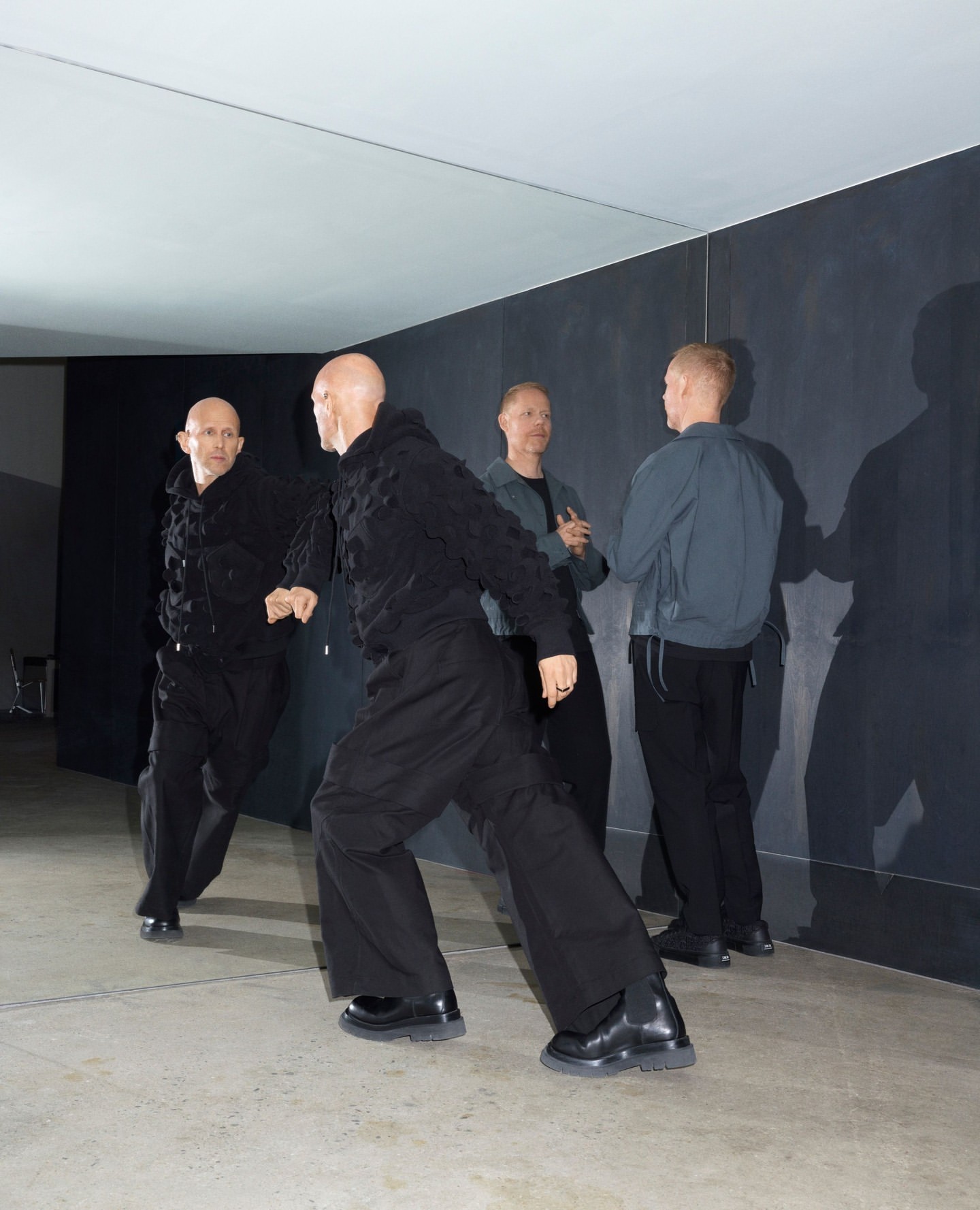 Four bald adult men indoors wearing dark clothing. Two of the men are facing forward, and the other two are standing behind them.
