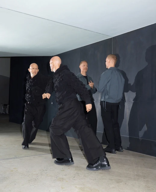 Four bald adult men indoors wearing dark clothing. Two of the men are facing forward, and the other two are standing behind them.