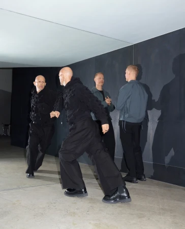 Four bald adult men indoors wearing dark clothing. Two of the men are facing forward, and the other two are standing behind them.