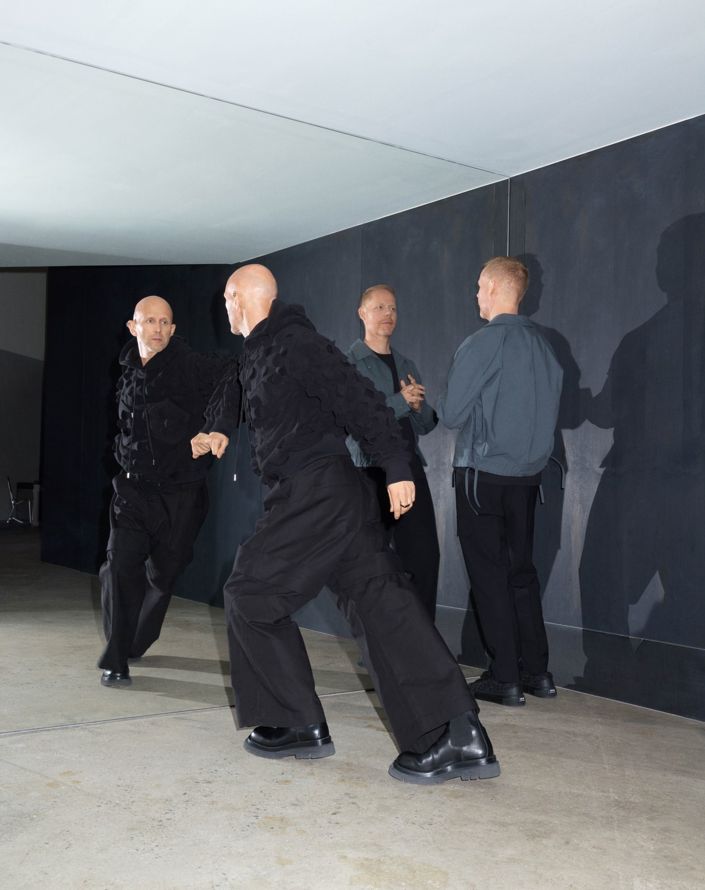 Four bald adult men stand in a minimalist interior, two near a mirror wearing dark clothing, while the others are in grey and black.