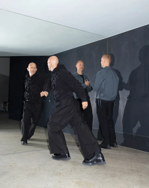 Four bald adult men stand in a minimalist interior, two near a mirror wearing dark clothing, while the others are in grey and black.
