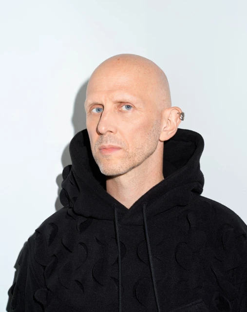 Close-up portrait of a bald adult male wearing a black patterned hoodie, against a white backdrop. The focus is on his face and attire.