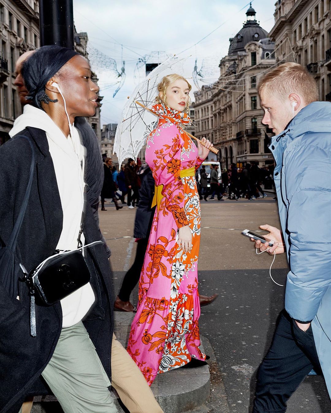 Street-style fashion shot of a blonde woman with a white parasol in a vibrant floral gown, walking among pedestrians in an urban setting.