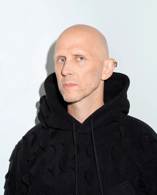 Portrait of a bald adult male with an ear piercing, wearing a black hoodie with textured design, set against a plain white background.