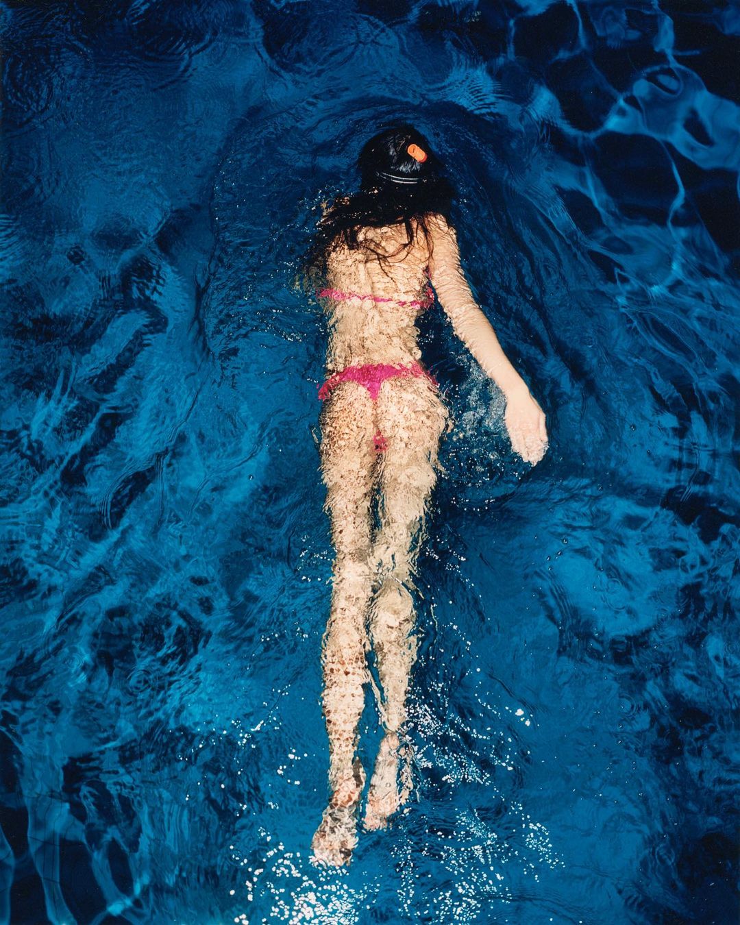 Overhead shot of a woman in a pink bikini and black swim cap swimming in a pool, with deep blue water and rippling light patterns.