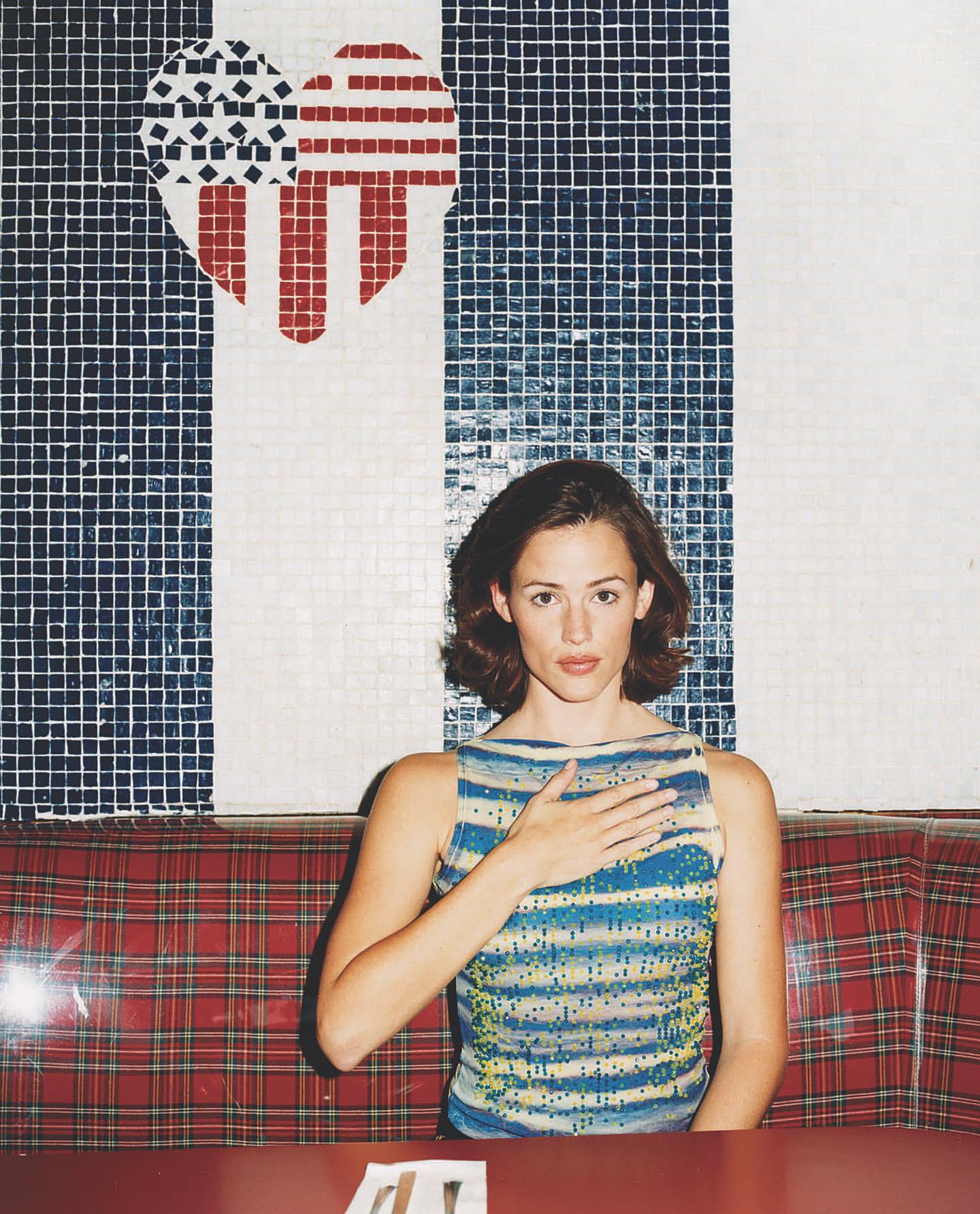 Jennifer Garner in a blue striped top with hand over her chest sitting in a red plaid booth, with a heart-shaped American flag mosaic.
