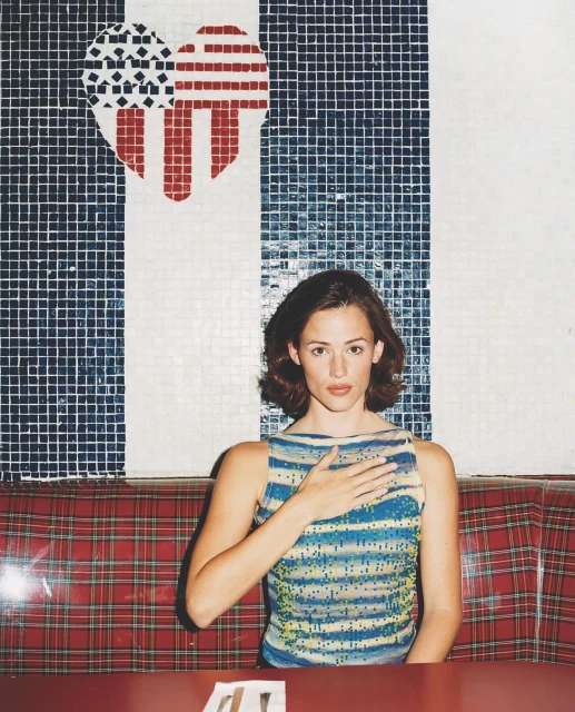 Jennifer Garner in a blue striped top with hand over her chest sitting in a red plaid booth, with a heart-shaped American flag mosaic.