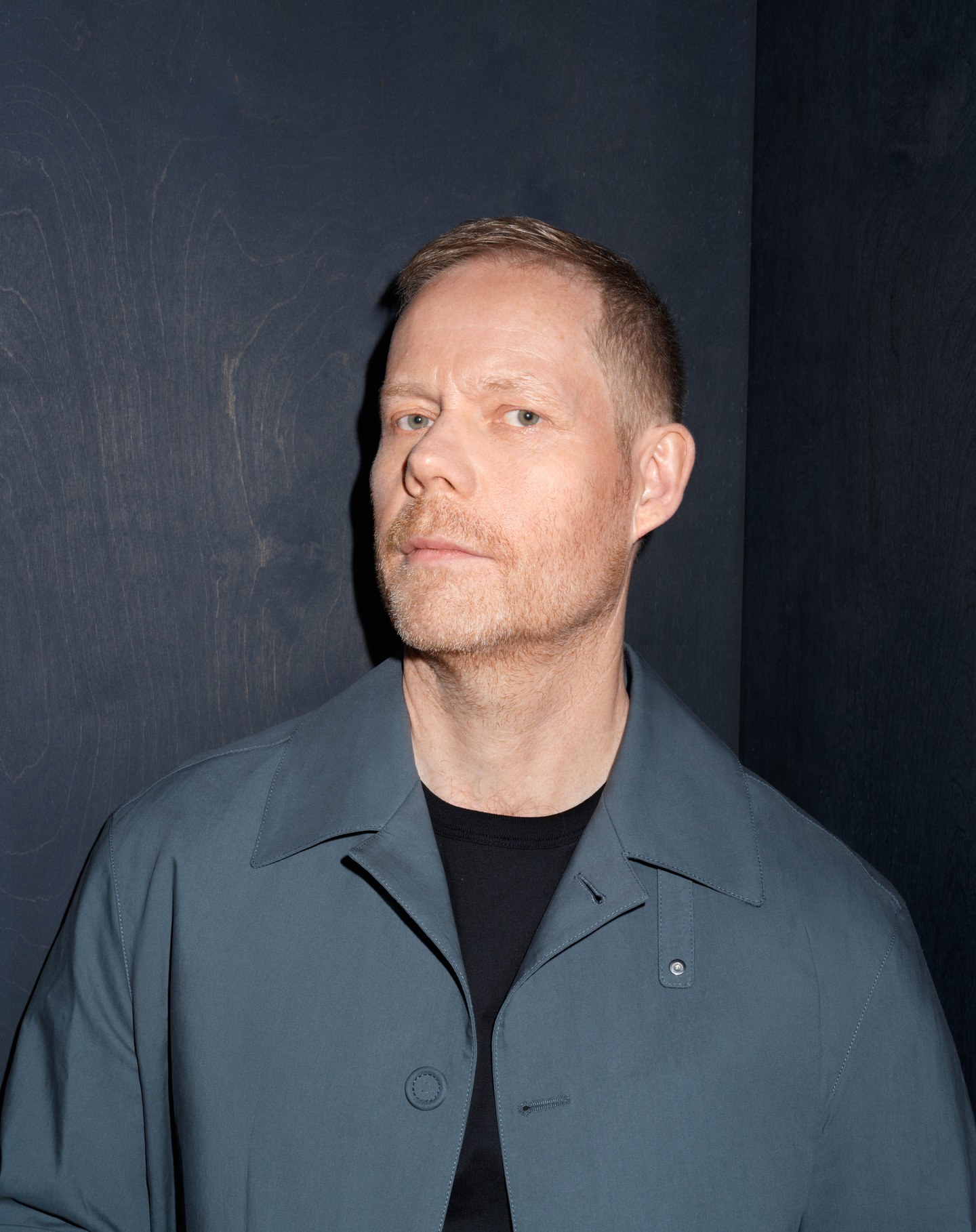 Portrait of a fair-skinned adult male wearing a teal jacket and a black t-shirt, standing indoors against a dark grey textured wall.