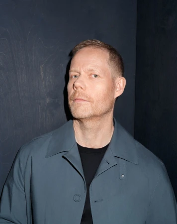 Portrait of a fair-skinned adult male wearing a teal jacket and a black t-shirt, standing indoors against a dark grey textured wall.