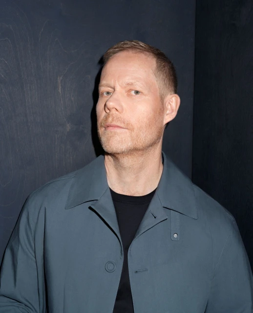Portrait of a light-skinned adult male with short hair wearing a blue-grey coat over a black shirt against a dark blue panelled background.