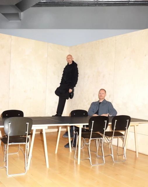 Two adult men pose indoors near a table and chairs. One man leans against a plywood wall, while the other sits at the table. 